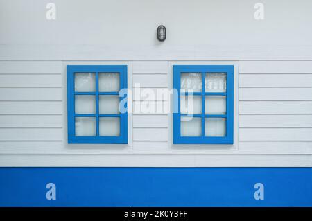 Zwei Fenster zur weißen Holzwand, Blick auf den Außenbereich. Vor dem Haus gibt es ein farbenfroh blaues Fenster. Stockfoto