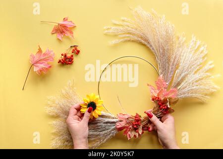 Hände, die getrockneten Blumenkranz aus trockenem Pampagras und Herbstblättern machen. Flach auf weiß gelbem Tisch, Sonnenlicht mit langen Schatten. Stockfoto