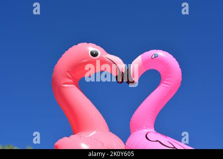 Zwei aufblasbare Flamingos aus Kunststoff, die ein Liebesherz gegen den blauen Himmel Formen Stockfoto