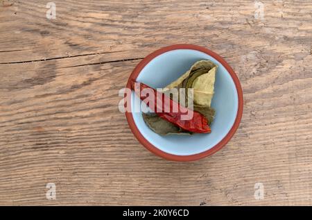 Draufsicht auf Kaffir-Limettenblätter und eine rote Chilischote in einer kleinen Schüssel isoliert auf einem Holzhintergrund. Stockfoto