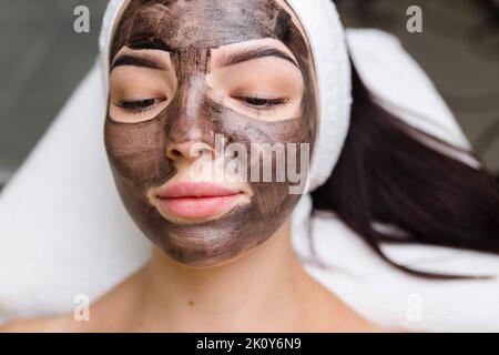 Kosmetologe, der schwarze Maske auf hübsches Frauengesicht mit schwarzen Handschuhen aufträgt, wunderschöne Frau im Spa, die Gesichtsbehandlungen hat Stockfoto