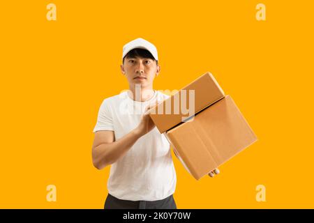 Junger asiatischer Lieferer mit Mütze und weißem, blanken T-Shirt, der Paketpostkasten auf gelbem Hintergrund isoliert hält. express-Zustellservice-Konzept. Stockfoto