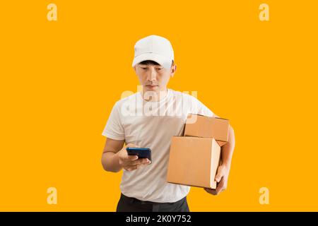 Junge asiatische Lieferung Mann trägt Kappe mit weißen leeren T-Shirt hält Smartphone und Paket-Briefkasten isoliert auf gelbem Hintergrund. express-Lieferung s Stockfoto
