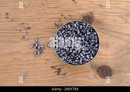 Draufsicht auf eine offene Dose Luftgewehrkugeln auf einer alten Holzplatte mit mehreren Pellets zur Seite. Stockfoto