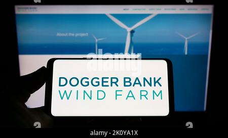 Person, die das Smartphone mit dem Logo des Offshore-Energiegenerators Dogger Bank Wind Farm auf dem Bildschirm vor der Website hält. Konzentrieren Sie sich auf die Telefonanzeige. Stockfoto