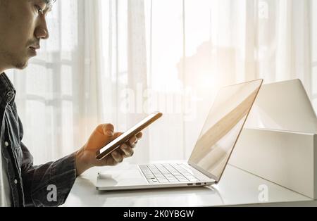 Die Hände des Mannes mit Smartphone und Laptop-Computer auf weißem Tisch zu Hause. Online-Shopping, Heimarbeit, Freiberufler, Online-Lernkonzept. Stockfoto