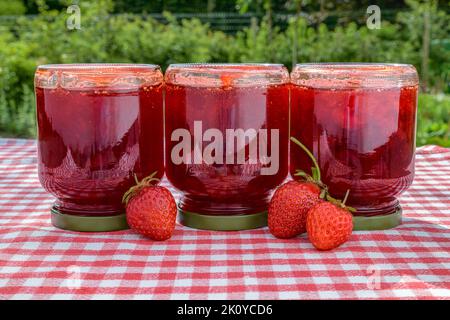 Eine Reihe von drei umgedrehten Gläsern mit frischer hausgemachter Erdbeermarmelade und drei Erdbeeren auf einem Tisch mit einer rot karierten Tischdecke. Stockfoto