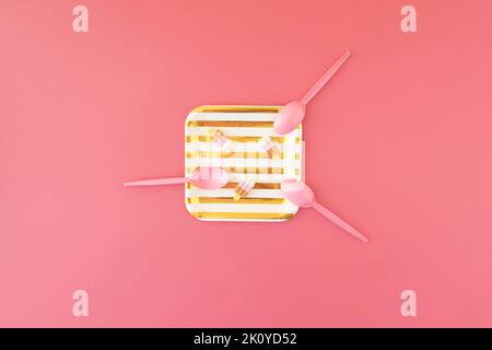 Gold- und weiß gestreifter Pappteller mit drei Gummieis und drei rosa Plastiklöffeln auf rosa Hintergrund. Stockfoto