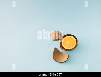 Minimalistisches Konzept mit einer halben frischen Orange in einer gebrochenen Kokosnussschale auf blauem Hintergrund. Stockfoto