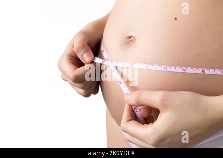 Junge Schwangerin Frau, die den wachsenden Bauch mit einem kaiserlichen Systemband misst, niemand, beschnitten, weißer Hintergrund Stockfoto