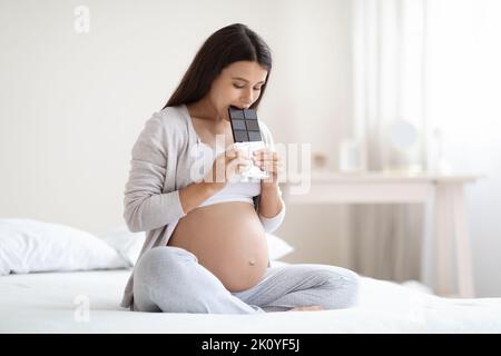 Junge Schwangere, die auf dem Bett sitzt und Schokolade isst Stockfoto