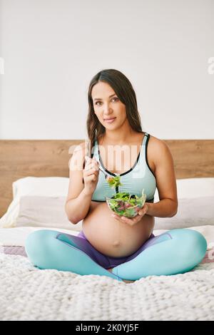 Porträt einer jungen Schwangeren in Sportkleidung, die im Bett einen frischen Salat isst. Bewusste Mutterschaft. Gesundes Lebensmittelkonzept Stockfoto