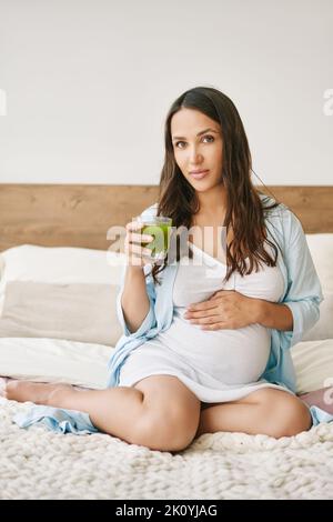 Porträt einer jungen Schwangeren im Pyjama, die grünen Smoothie Detox-Saft im Bett trinkt. Bewusste Mutterschaft. Gesundes Lifestyle-Konzept Stockfoto