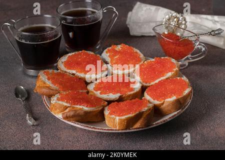 Hausgemachte Sandwiches mit rotem Kaviar auf Weißbrot-Toasts auf runden Teller und zwei Tassen Kaffee auf braunem Hintergrund Stockfoto