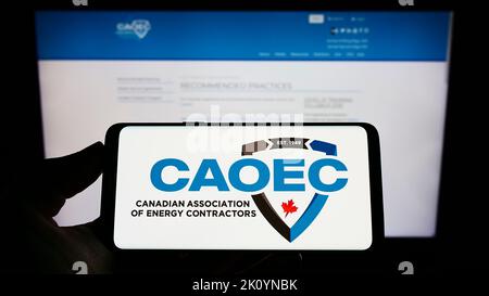 Person mit Mobiltelefon und Logo der Canadian Association of Energy Contractors (CAOEC) auf dem Bildschirm vor der Webseite. Konzentrieren Sie sich auf die Telefonanzeige. Stockfoto