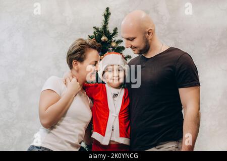 Mutter, Vater und Sohn im Weihnachtsmann-Kostüm stehen in ihrer Wohnung in der Nähe des Weihnachtsbaums, Eltern schauen auf ihren Sohn und lächeln. Stockfoto