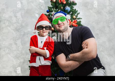 Ein Junge und sein Vater in lustigen Sonnenbrillen und Weihnachtsmützen stehen neben dem Weihnachtsbaum mit gekreuzten Armen auf der Brust. Spiele für Weihnachten Stockfoto