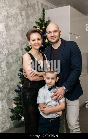 Familie - Mama, Papa und Sohn in smarter Kleidung sitzen neben dem Weihnachtsbaum in ihrer neuen Wohnung und schauen auf die Kamera. Stockfoto