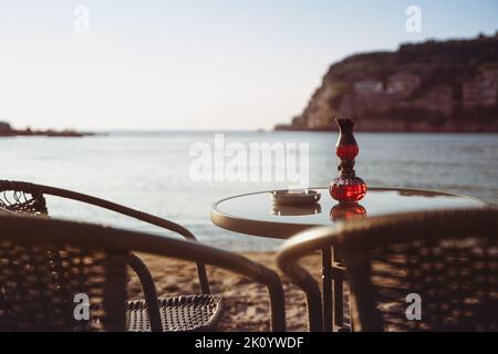 Rückansicht Nahaufnahme von leerem Kaffeetisch und Stühlen am Sandstrand mit Aschenbecher und Kerzenlicht vor dem Golf am Meer mit klarem Himmel vor Sonnenuntergang Stockfoto