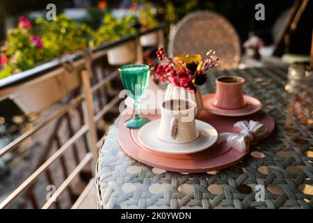 Vorderansicht Nahaufnahme der traditionellen türkischen Kaffeetassen, die draußen mit Wasserblumen serviert werden, und das Dessert in romantischer Umgebung auf einem Glastisch bei Sonnenuntergang Stockfoto