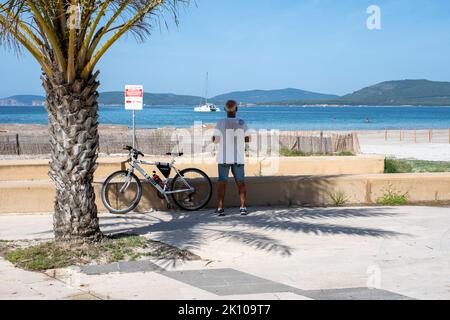 Mann mit Fahrrad, der die Yacht von der Promenade Lungomare auf der Via Lido von Spiaggia del Lido in Alghero, Sardinien, Italien aus betrachtet Stockfoto