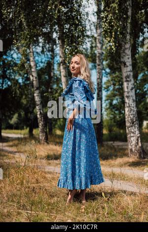 Eine junge Frau in einem langen Kleid steht an einem sonnigen Sommertag auf einer Lichtung. Stockfoto