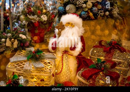 Eine Weihnachtsmann-Puppe im roten Pelzmantel und Hut mit Stab und Bart und Schachteln mit Weihnachtsgeschenken auf dem Hintergrund der Weihnachtsdekoration. Neu Ja Stockfoto