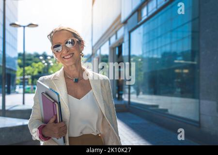 Kaukasische Geschäftsfrau mittleren Alters mit Blick auf die Kamera. Schöne reife Dame lächelt mit Sonnenbrille, Büchern und Tablet. Stockfoto