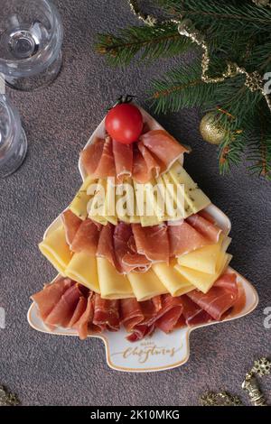 Vorspeise mit Schinken und Käse in baumförmiger weihnachtlicher Platte und Gläsern auf braunem Tisch Stockfoto