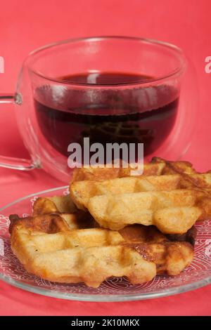 Flauschige Waffeln. Sie liegen auf einer Untertasse. Neben einer Tasse Kaffee. Der Hintergrund ist rosa. Nahaufnahme. Stockfoto