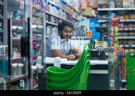 Junger männlicher pakistanischer Kassierer in einem 24-Stunden-Supermarkt in Dubai, Vereinigte Arabische Emirate. Der Mann sitzt mit beiden Armen auf dem Ladentisch und lächelt der Kamera zu. Stockfoto