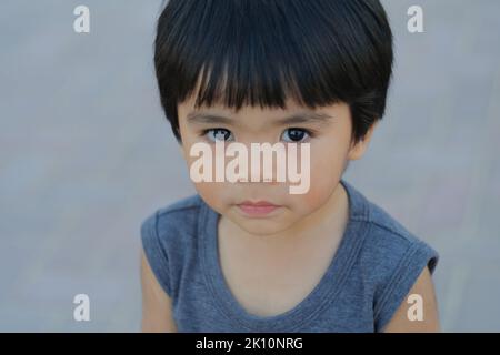 Netter philippinischer Junge, etwa 5 Jahre alt, schaut vor die Kamera. Nahaufnahme eines Tages im Freien mit einem jungen asiatischen Kind mit glänzendem, glattem Haar und schwarzen Augen. Stockfoto