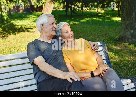 Hübsches, multirassisches, älteres, graues Haar-Paar, das sich nach einem Spaziergang auf der Bank im Park ausruht, am Morgen lächelnd auf der Bank sitzt und im sonnigen Sommer Entspannung im Freien genießt Stockfoto