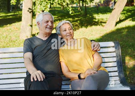 Glückliches, multirassisches, älteres, graues Haar-Paar, das sich nach einem Spaziergang auf der Bank im Park ausruht, am Morgen lächelnd auf der Bank sitzen und im sonnigen Sommer im Freien entspannen Stockfoto