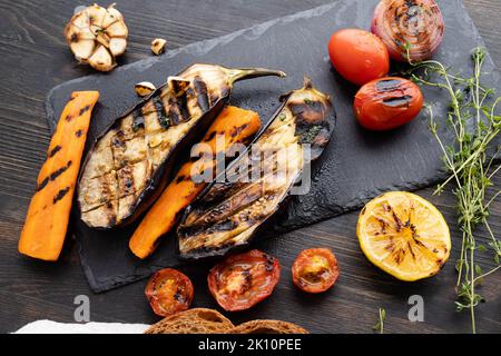 Gegrilltes Gemüse mit Kräutern: Tomaten, Auberginen, Zwiebeln, Karotten, Knoblauch, Brot auf einem dunklen Holzhintergrund auf einem Schneidebrett. Speicherplatz kopieren. Flach liegend. Lifestyle. Stockfoto