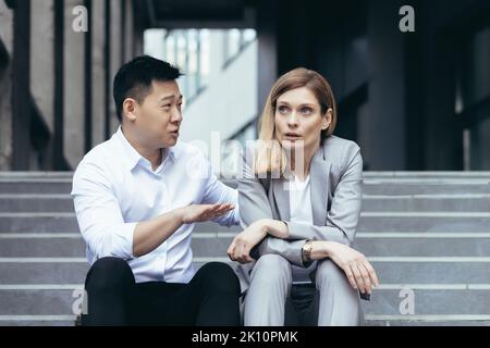 Interrassischer Konflikt am Arbeitsplatz. Eine junge Frau und ein junger asiatischer Mann sitzen auf den Stufen vor einem Büro und streiten sich. Ein Mann schreit und beweist einer Frau etwas. Die Frau hört erittlich zu. Stockfoto