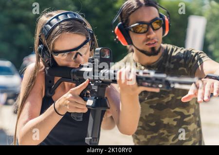 Langhaariges Mädchen übt das Zielschießen mit einem Instruktor und hält ein schwarzes Gewehr mit einem Kollimator auf dem Schießstand, mittlere Nahaufnahme. Hochwertige Fotos Stockfoto