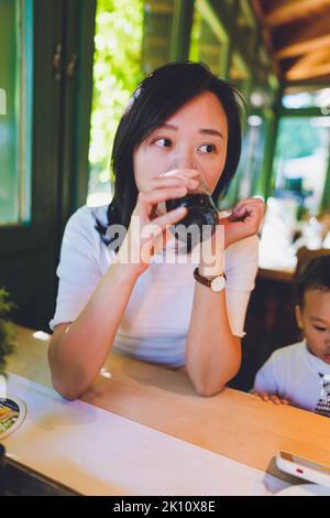 Eine Frau, die in einem Restaurant sitzt und trinkt. Ihr Kind sitzt neben ihr. Stockfoto