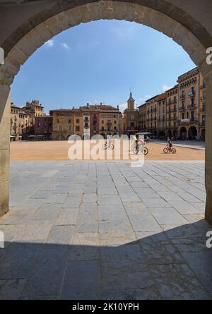 VIC, Spanien - 11. September 2022: Plaza de Vic mit einigen Radfahrern in Katalonien, Spanien, durch einen der Bögen der umliegenden Säulenhallen gesehen. Stockfoto