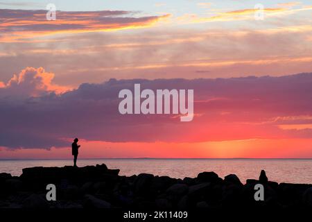 Dramatische Szene an der Küste von Neuengland. Dunkle Silhouetten zweier unverkennbarer Menschen auf Wellenfelsen am Cape Cod Beach, die den farbenfrohen Sonnenuntergang betrachten. Stockfoto