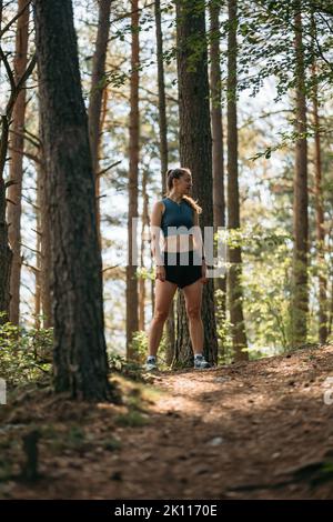 Die junge Läuferin in Sportkleidung steht auf dem Trail im grünen Wald, bereit zum Laufen, während sie sich auf den Marathon vorbereitet Stockfoto