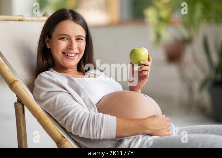 Gesunder Snack. Schöne Schwangerin sitzt im Stuhl und hält grünen Apfel Stockfoto