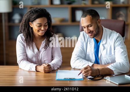 Fröhlicher afroamerikanischer Arzt und Patient, der am Tisch sitzt Stockfoto