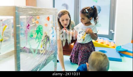 Frischhaltefolie für die Verbesserung der Phantasie von Kindern. Kleinkinder malen mit, Bürsten, Rollen und Farben auf einer Frischhaltefolie, die um den hölzernen Regalständer gewickelt ist. Lustige Aktivität für Kinder Sinnesfertigkeiten, Kreativität, räumliche Aufmerksamkeit und visuelle Wahrnehmungsentwicklung im Kindergarten. Hochwertige Fotos Stockfoto