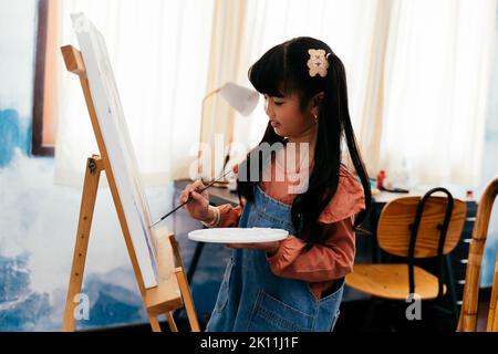 Nettes asiatisches Mädchen in legerer Kleidung mit Zöpfen, die Palette halten und Bild auf Leinwand malen, während sie in der Nähe von Staffelei im hellen Raum zu Hause stehen Stockfoto