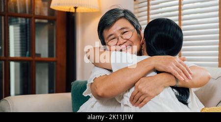 Glückliche asiatische Tochter lächelt mit geschlossenen Augen und umarmt ältere Mutter, während sie am Wochenende im Wohnzimmer zu Hause auf der Couch sitzt Stockfoto
