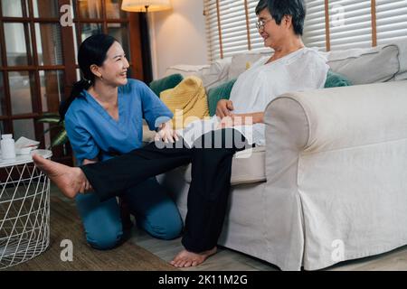 Glückliche asiatische Betreuerin lächelnd und streckend Bein einer älteren Frau auf dem Sofa während der Rehabilitation Sitzung im hellen Raum zu Hause sitzen Stockfoto