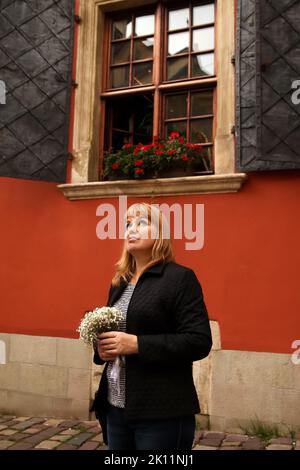 Unschärfe-Frau, die auf rotem Wandhintergrund steht und weiße Blumen hält. 40s seriöse Frau. Speicherplatz kopieren. Außen Porträt weiblich. Brautpaar. Aus Stockfoto