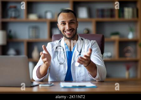 Fröhlicher afroamerikanischer Arzt, der in der Klinik am Schreibtisch sitzt und gestikuliert Stockfoto