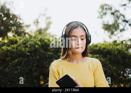 Teenagerin in gelbem Pullover, die im Park über Kopfhörer Musik auf ihrem Smartphone spielt Stockfoto